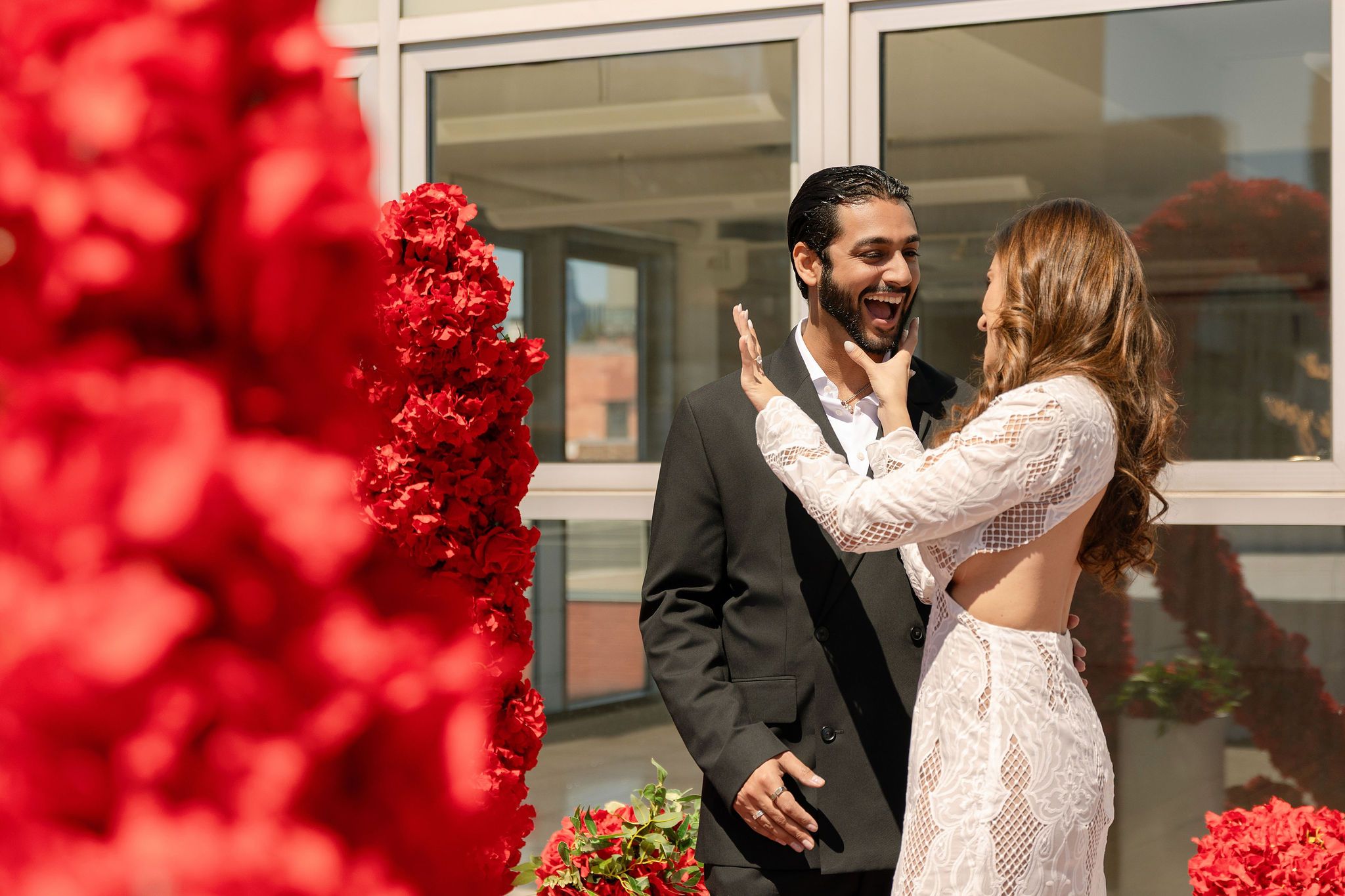 Couple in elegant wedding attire laughing together outdoors surrounded by vibrant red floral arrangements.