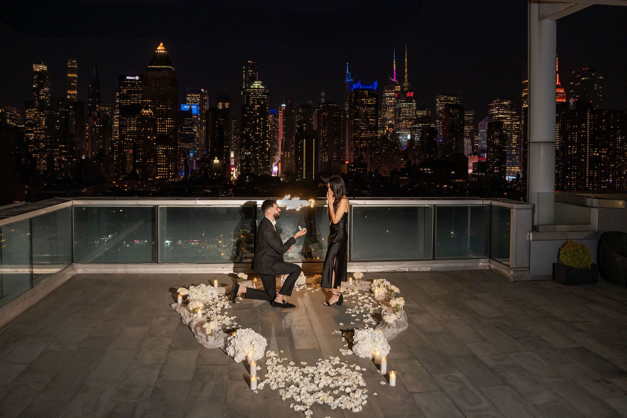 Romantic rooftop proposal with NYC skyline at night, man on one knee surrounded by white flowers and candles.