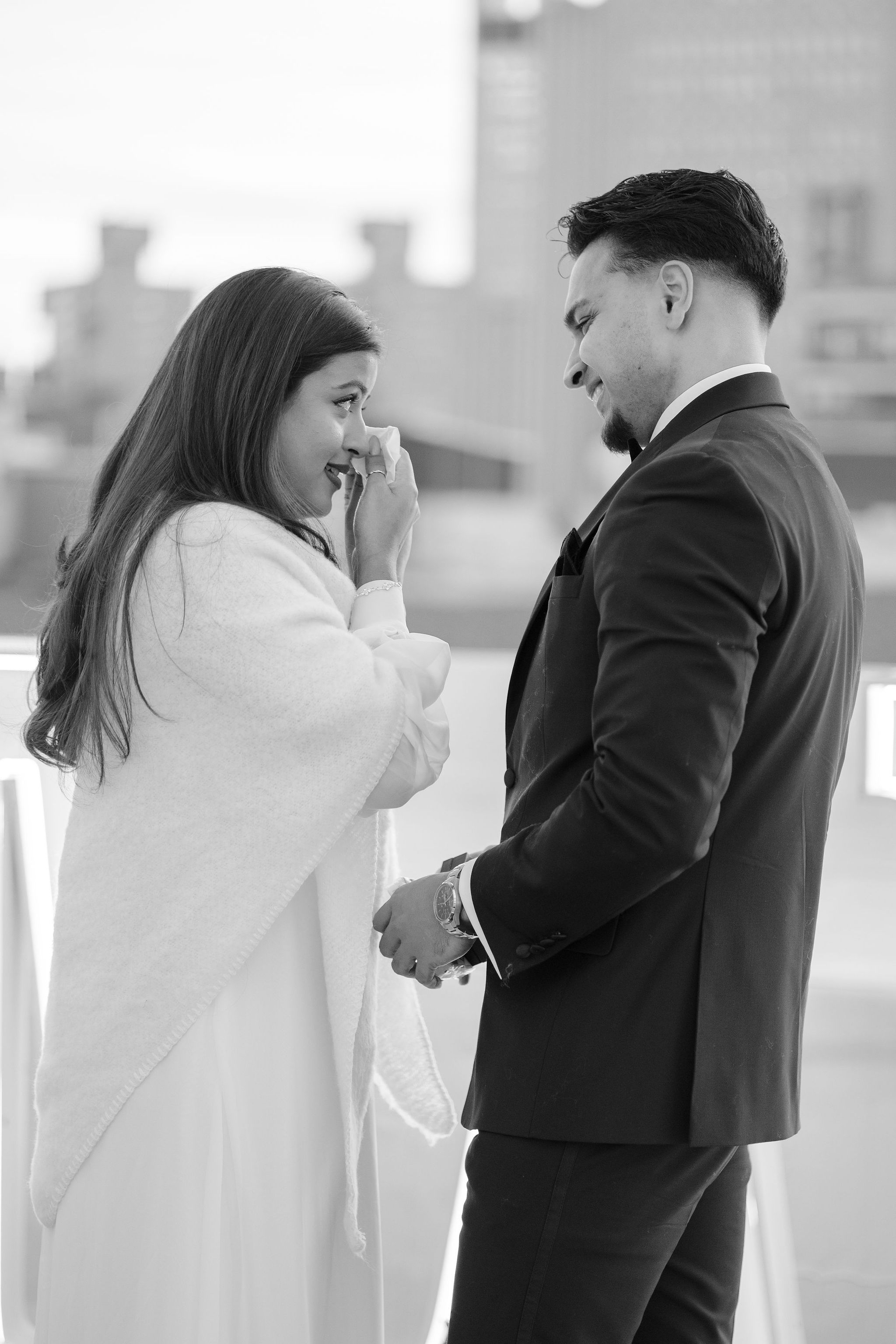 Black and white photo of emotional bride wiping tears while groom smiles holding hands during outdoor wedding ceremony.