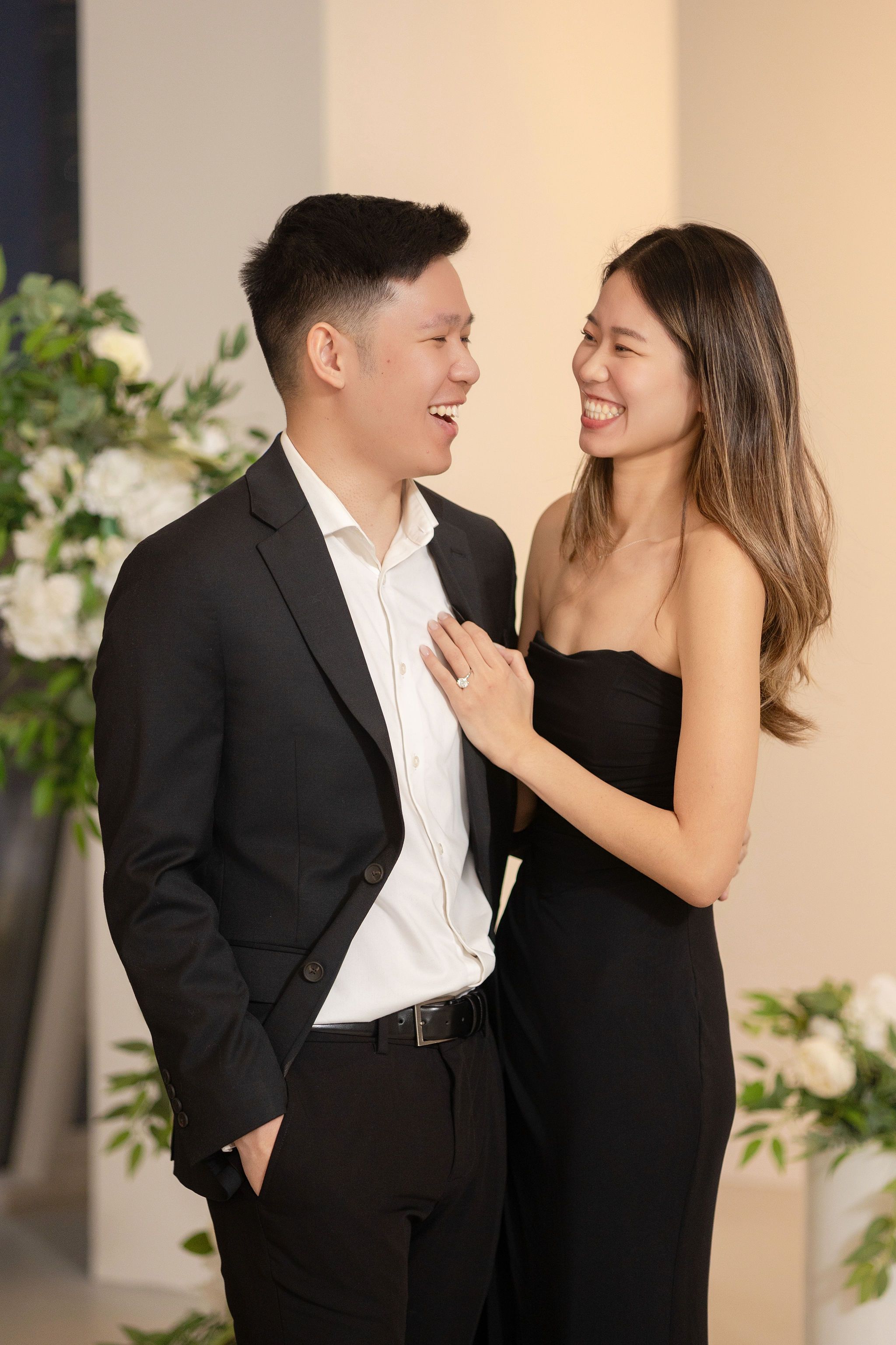 Smiling Asian couple in black formal attire sharing a joyful moment surrounded by white floral arrangements.