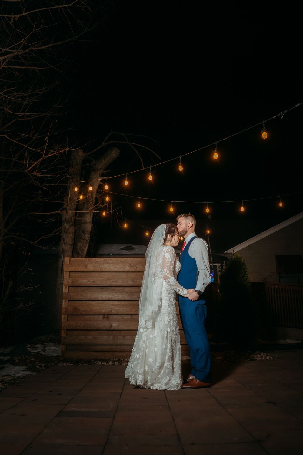 Bride and groom share a romantic moment under string lights at night, bride in lace gown with veil, groom in blue suit.