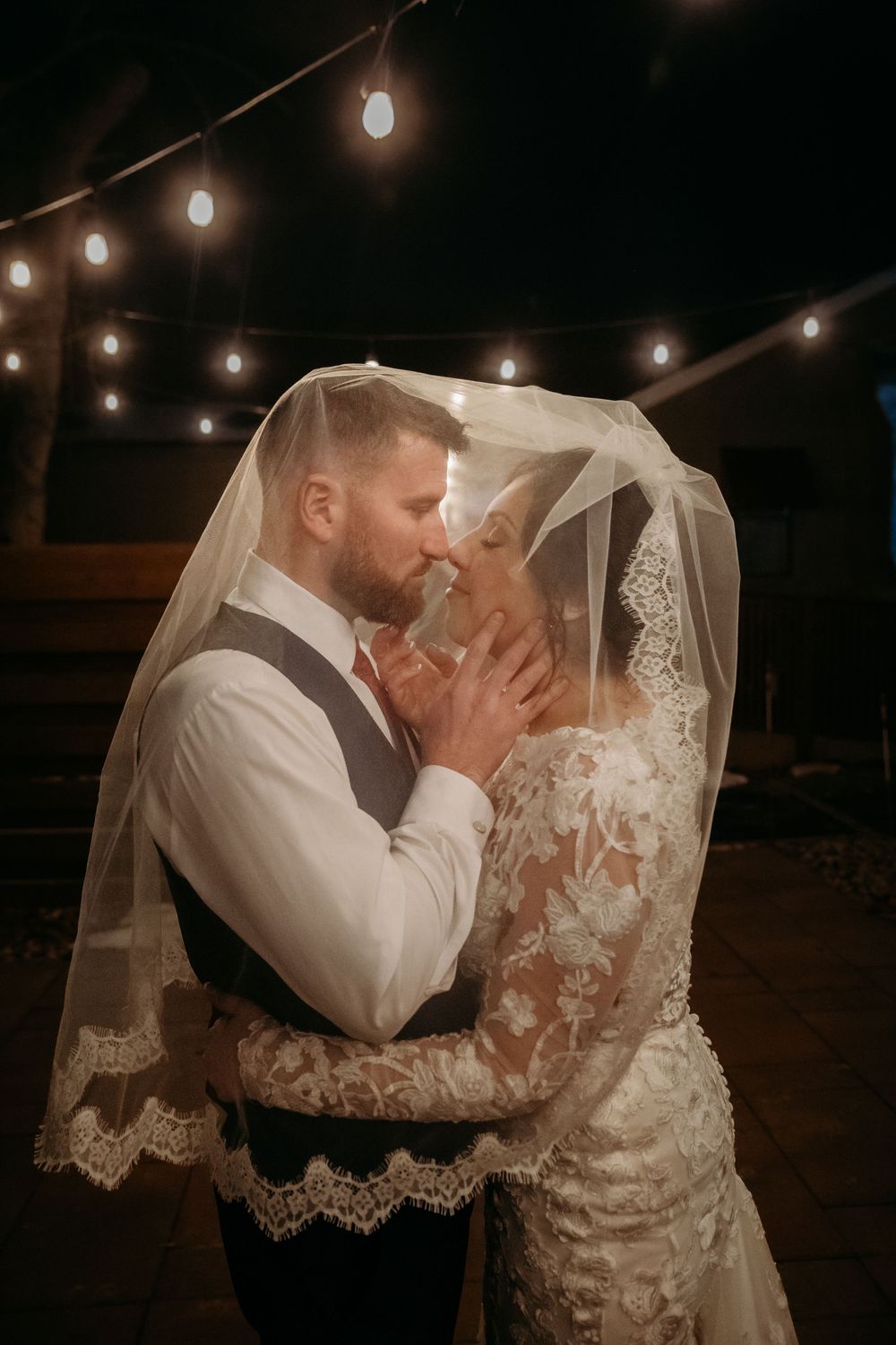 A bride and groom share an intimate moment under her lace veil, surrounded by warm string lights at their evening wedding reception.