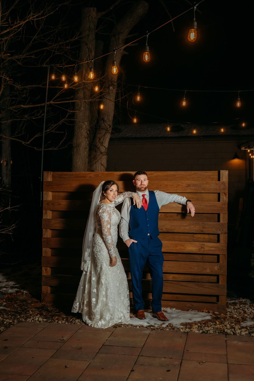 Bride in lace gown and groom in blue suit pose by wooden wall with string lights at night.