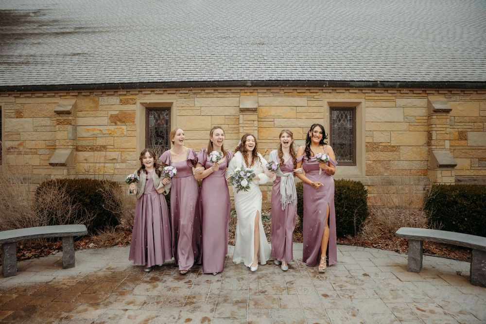 Bride in white gown poses with five bridesmaids in mauve dresses outside a stone building.