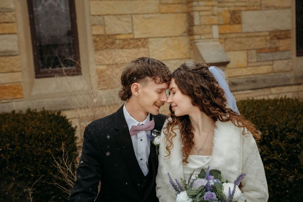 Newlywed couple sharing a romantic moment outdoors near a stone church, bride holding a purple bouquet.