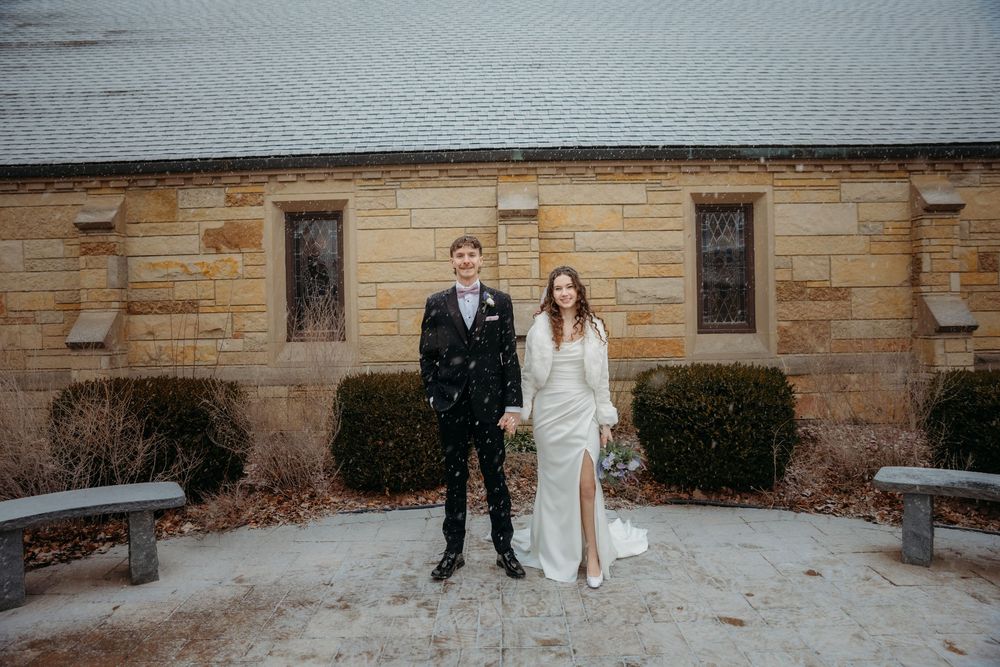 Bride and groom standing together in light snow outside a stone church building on their winter wedding day.