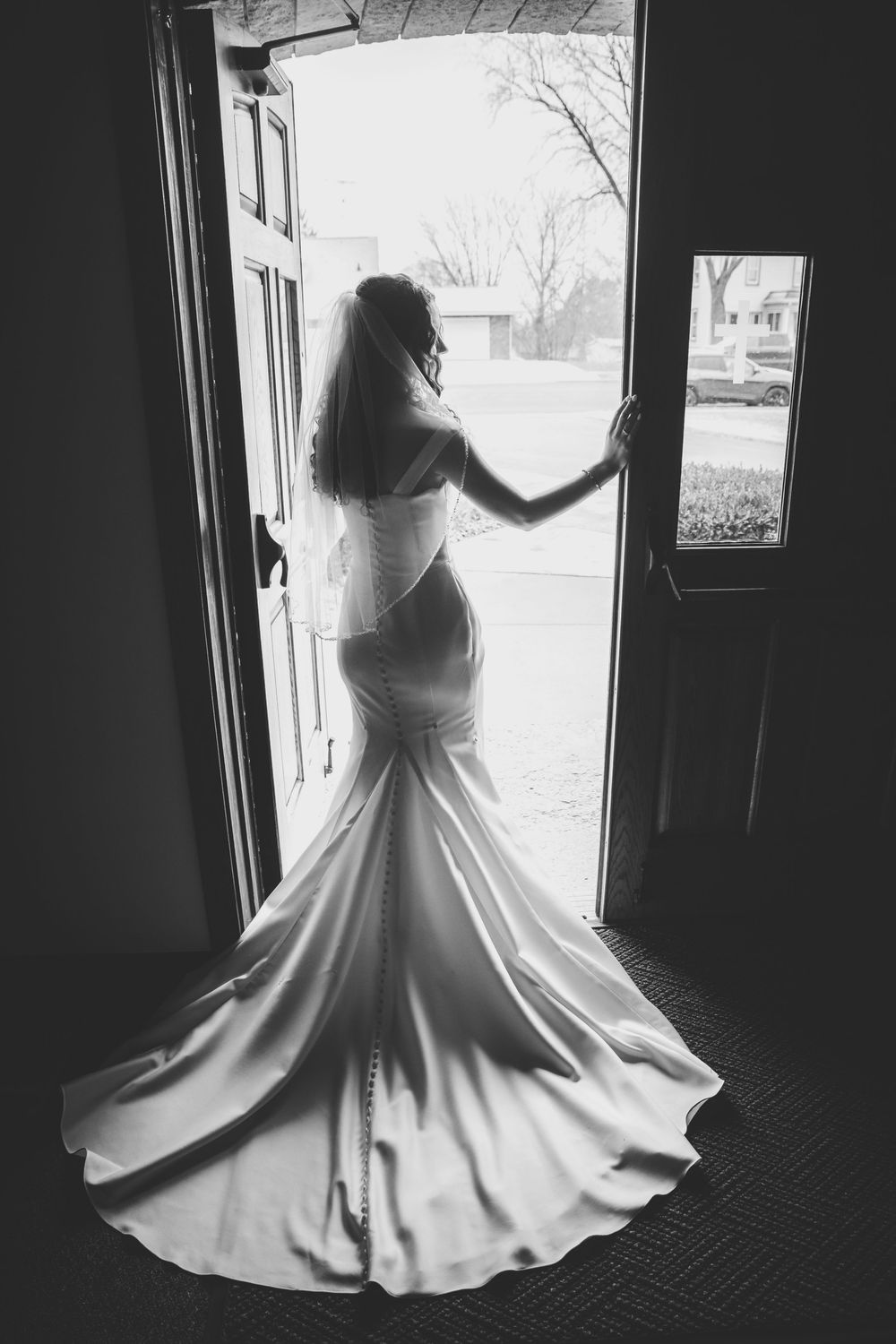 Black and white photo of a bride in a mermaid wedding gown with veil standing in an open doorway with train spread.
