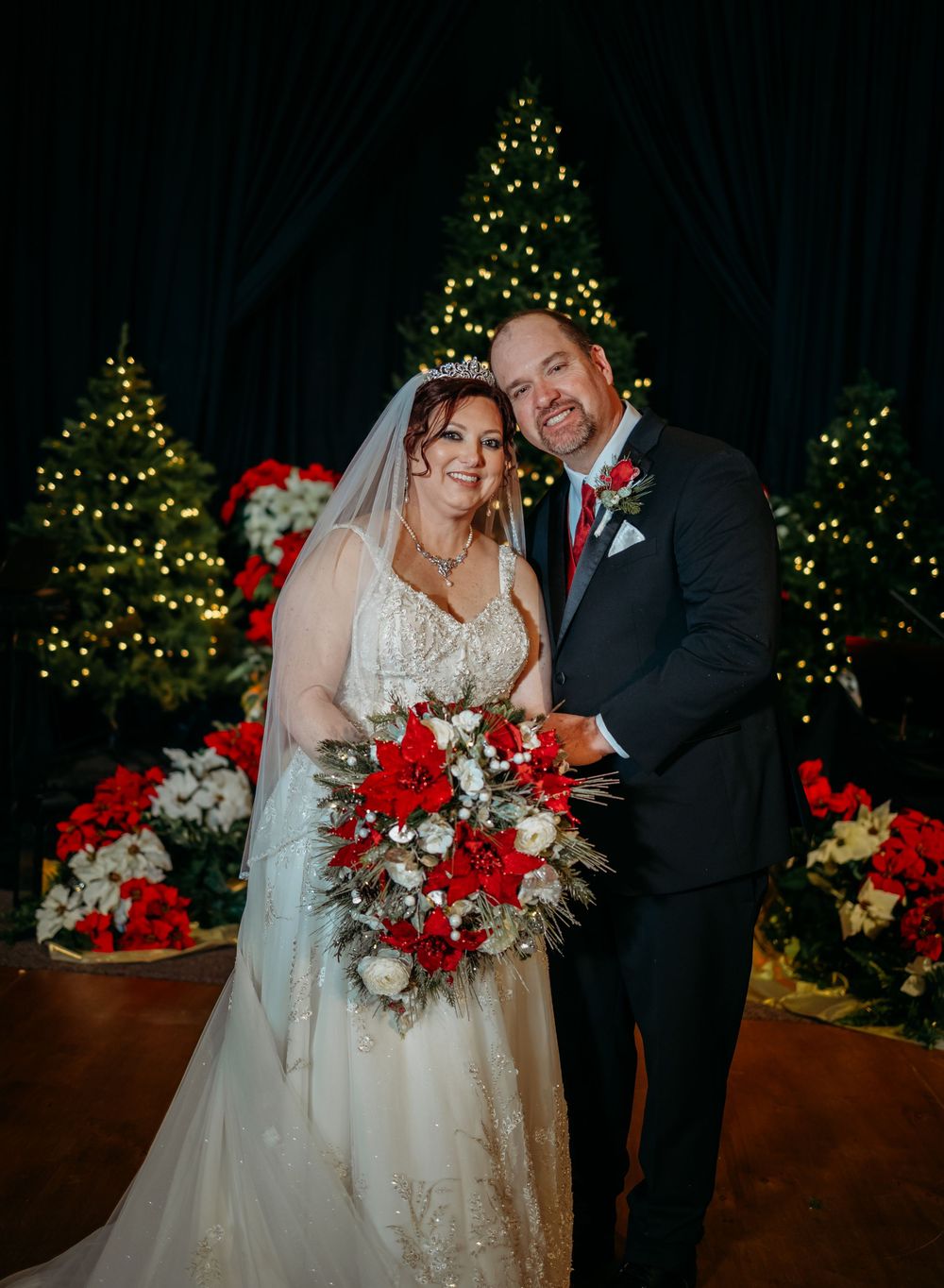 Bride and groom pose together at Christmas wedding with red poinsettias and lit trees in background.