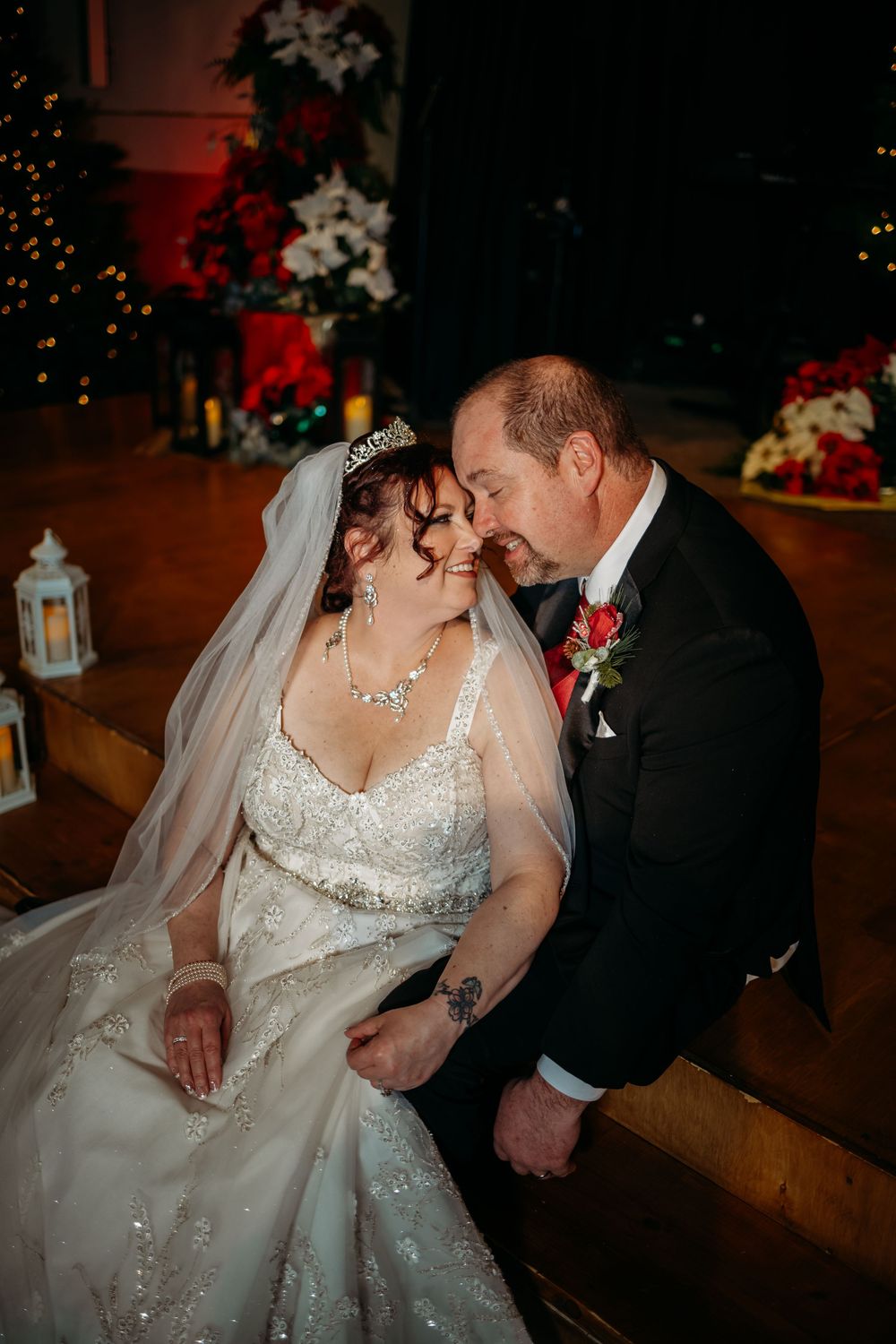 A bride and groom share an intimate moment seated in a church pew, adorned in wedding attire with holiday decorations in the background.
