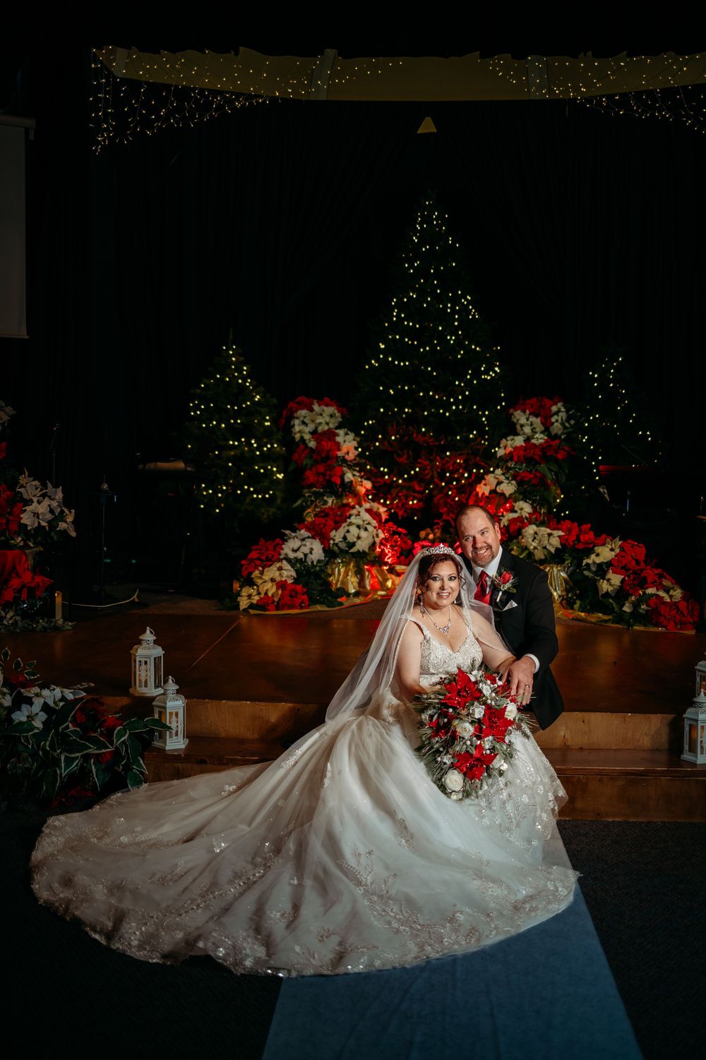 Bride and groom pose at Christmas wedding altar with poinsettias and lit trees in elegant church ceremony.