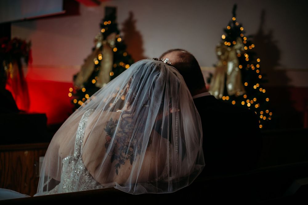 Bride and groom share a kiss under a wedding veil near Christmas trees with warm bokeh lights.