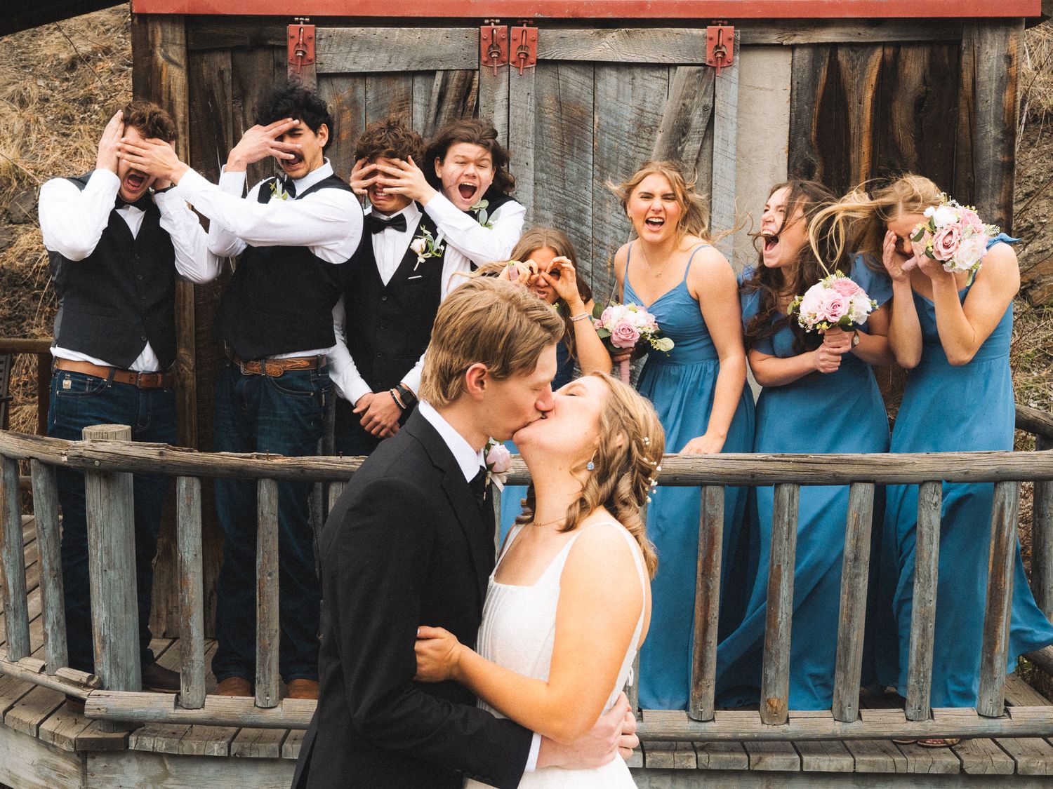 Bride and groom kissing while wedding party reacts with excitement on rustic wooden deck.