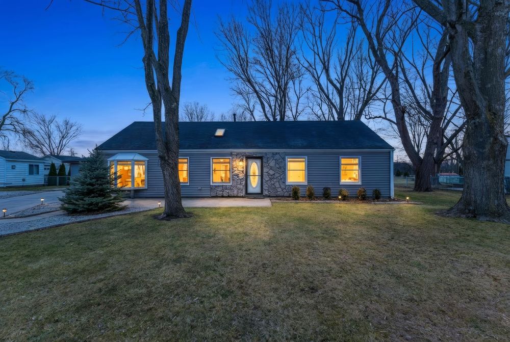 Charming ranch-style home with stone accents, glowing windows at dusk, surrounded by mature trees and a lush green lawn.