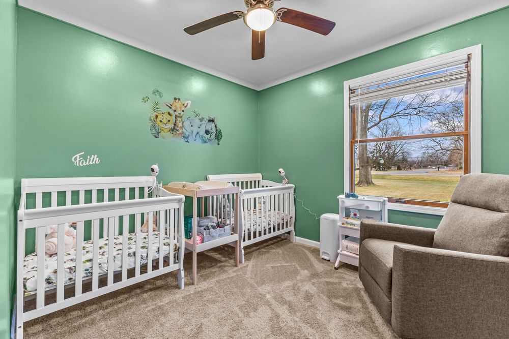 Green nursery with two white cribs, ceiling fan, carpeted floors, and natural light from a window overlooking yard.