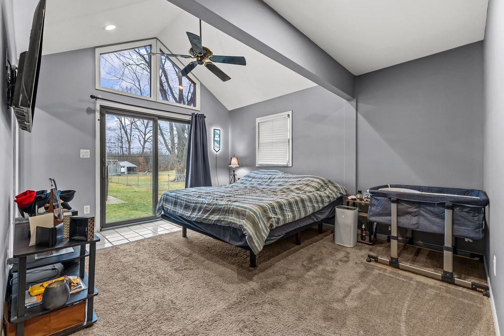 Spacious bedroom with vaulted ceiling, ceiling fan, plaid bedding, sliding glass door, and wall-mounted TV on gray walls.