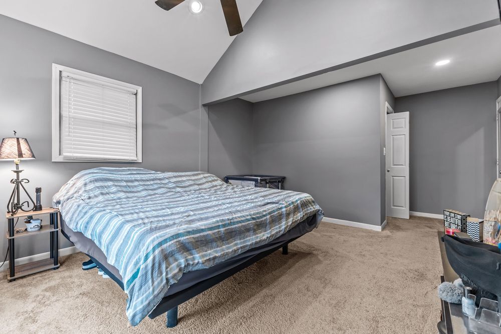 Spacious bedroom with vaulted ceiling, ceiling fan, plush carpet, and a queen bed with blue striped bedding near a window.
