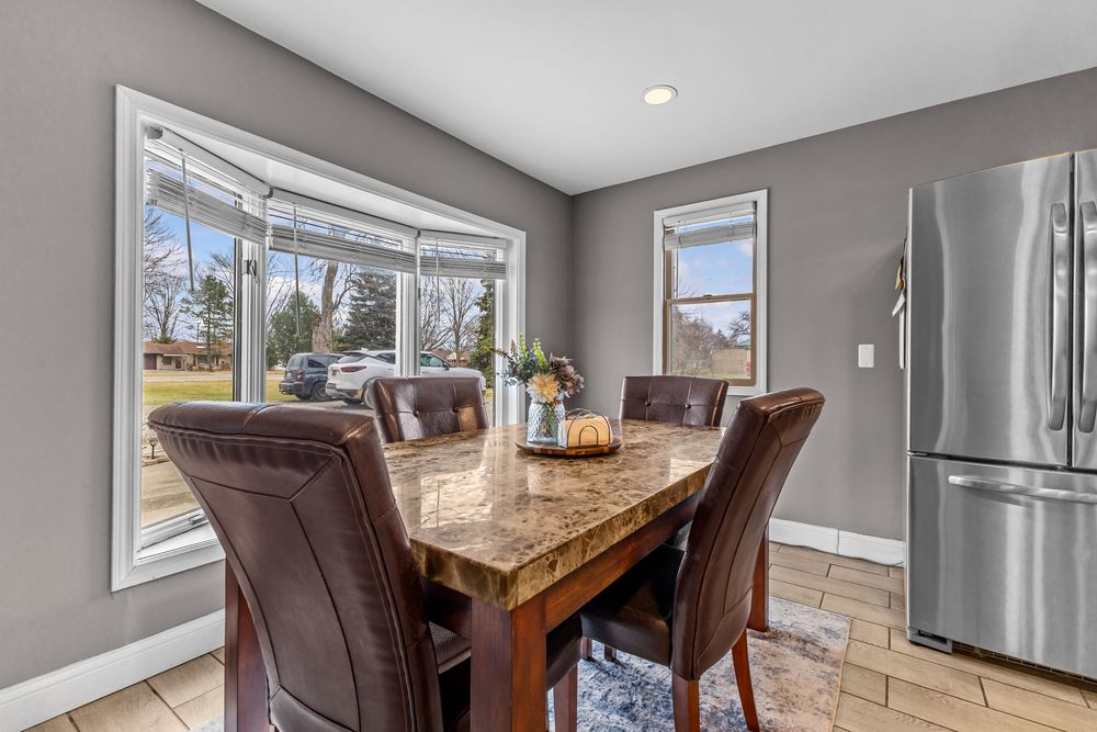 Bright dining area with marble-top table, brown leather chairs, bay window, and stainless steel refrigerator on tile floors.