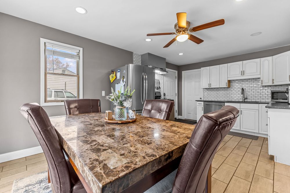 Modern dining area with granite table, brown leather chairs, white kitchen cabinets, subway tile backsplash, and ceiling fan.