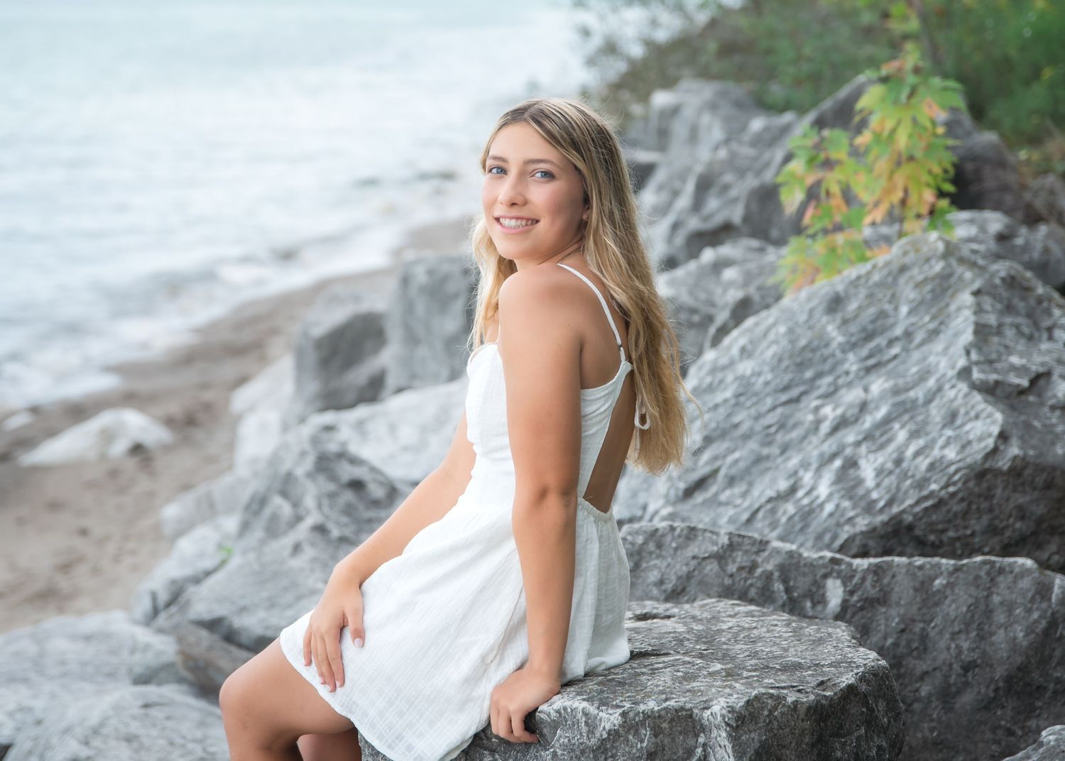 Young woman in white dress sitting on rocks by a lake shore, smiling over her shoulder on a sunny day.