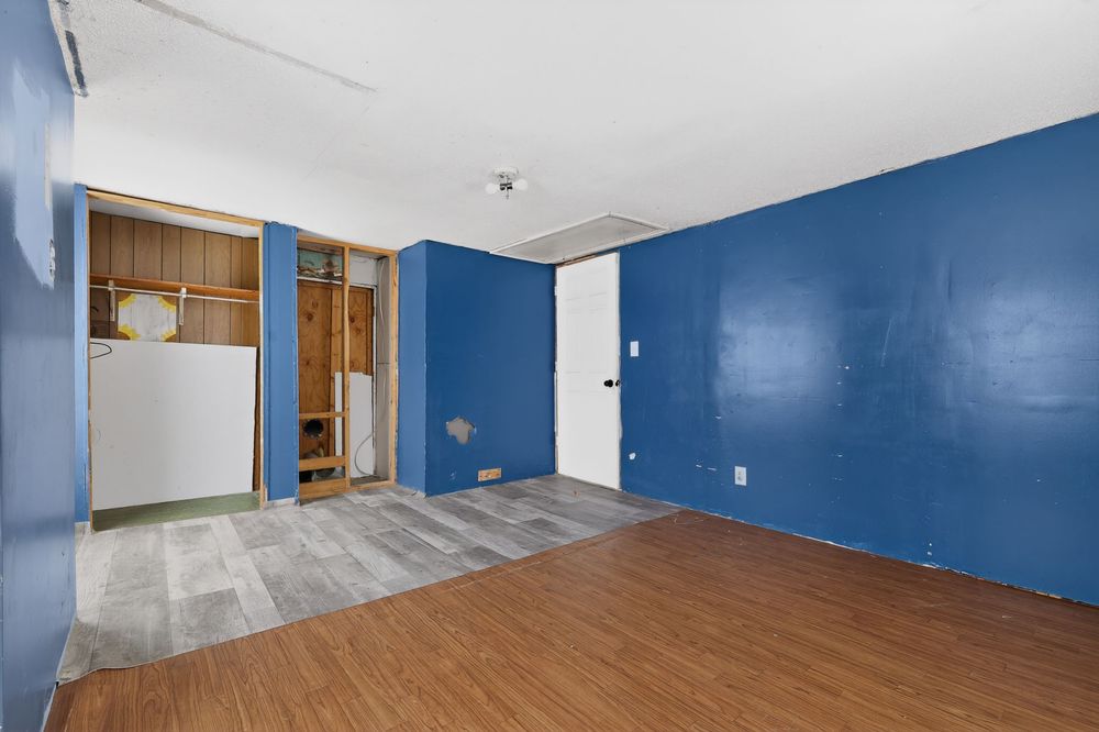 Empty room with blue accent walls and hardwood floors leading to a partially renovated space with open shelving.