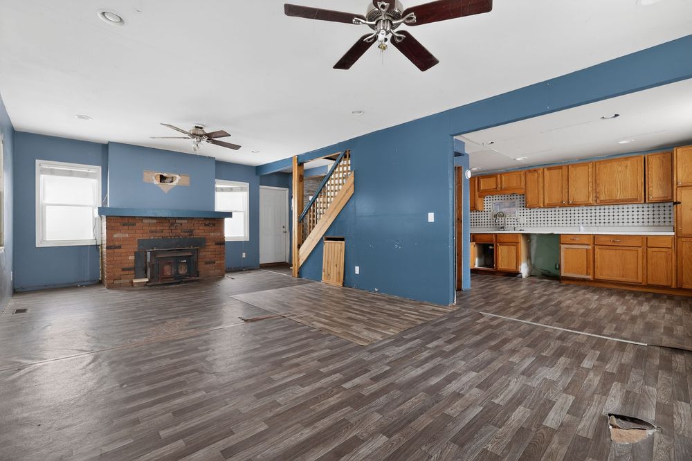 Open living room with blue walls, hardwood floors, brick fireplace, ceiling fan, and kitchen visible in background.