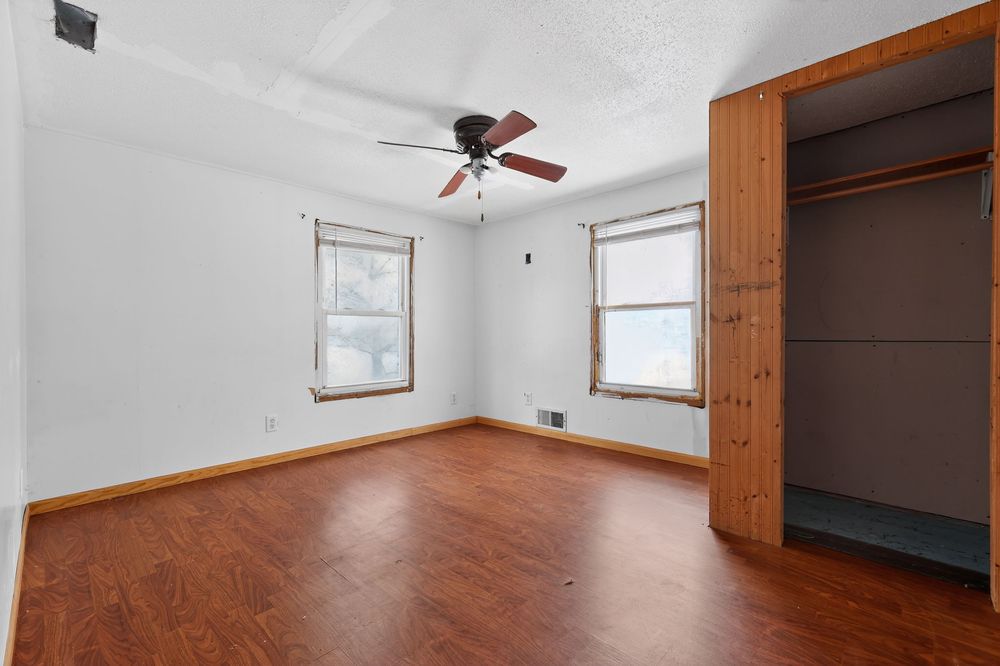 Bright bedroom with hardwood floors, ceiling fan, two windows, and an open closet with wood paneling.