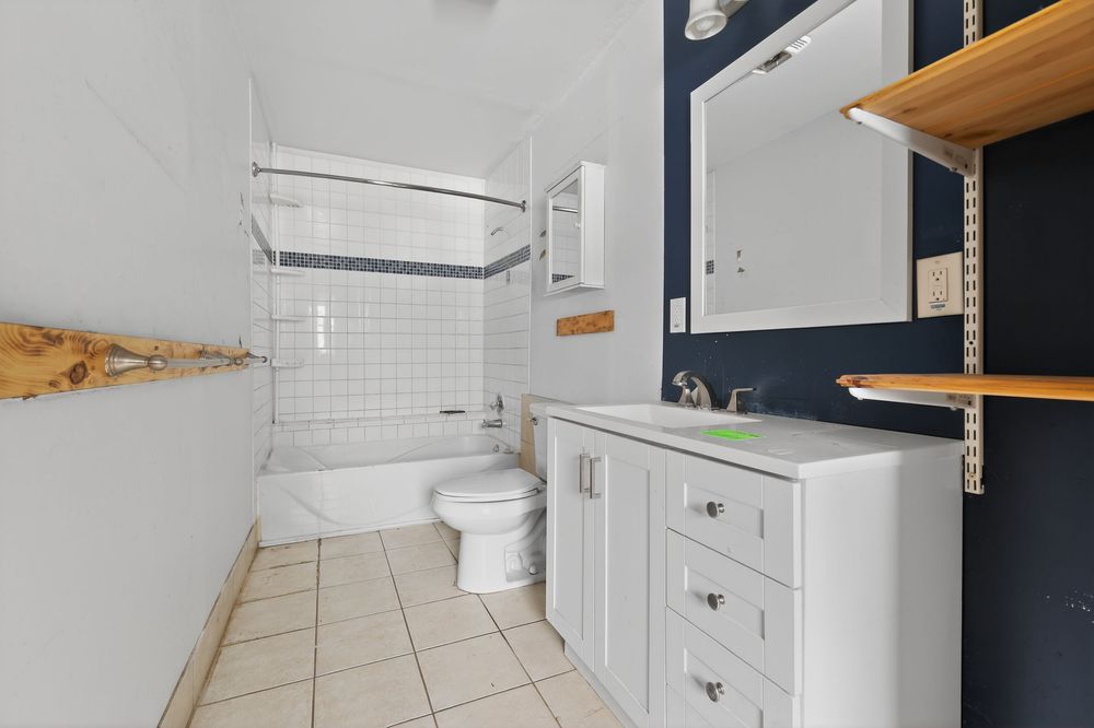 White bathroom with tiled shower-tub combo, toilet, white vanity with drawers, and navy blue accent wall with wood shelving.