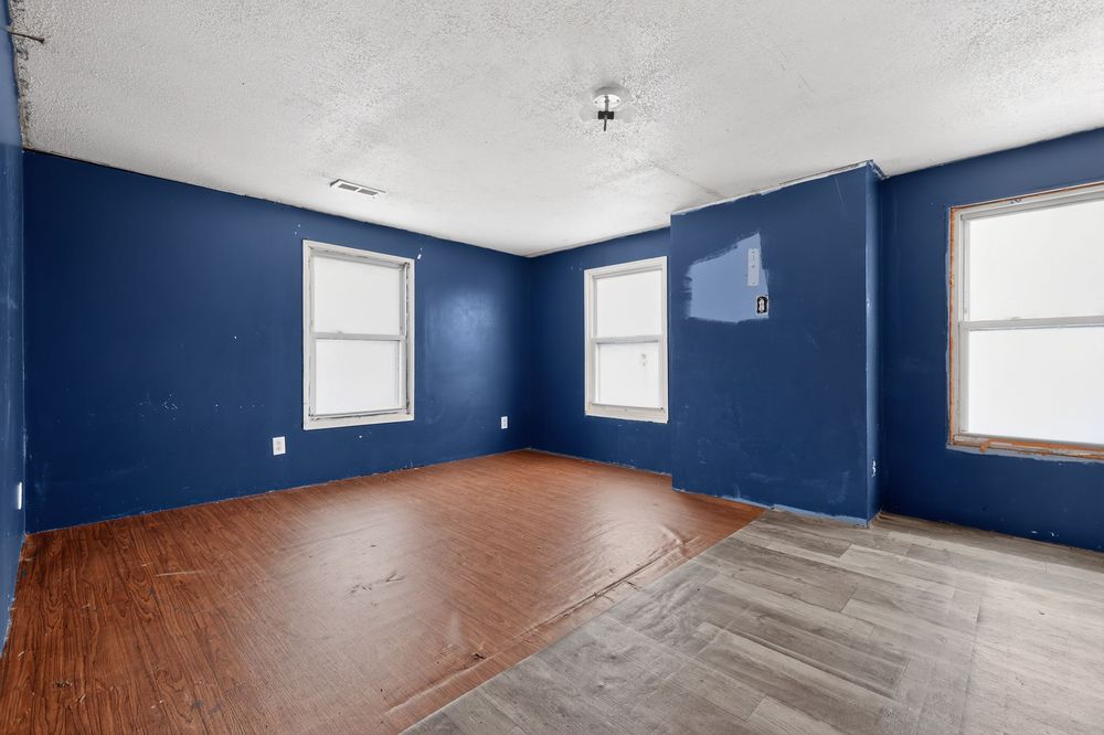 Empty room with bold navy blue walls, hardwood floors, and three windows allowing natural light in a fixer-upper home.
