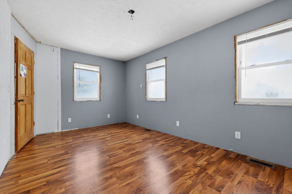 Empty room with hardwood floors, blue-gray walls, three windows, and a wooden door in a residential home.