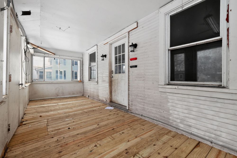 Enclosed porch with hardwood floors, white brick walls, and a white door with glass panels letting in natural light.