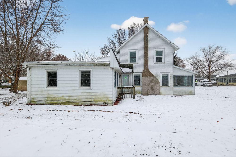 Snow-covered yard surrounds a white two-story farmhouse with a brick chimney and sunroom addition in winter.