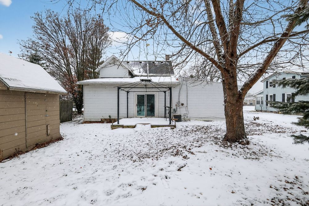 Snow-covered backyard of a white house with a large bare tree, small pergola, and adjacent garage in winter.