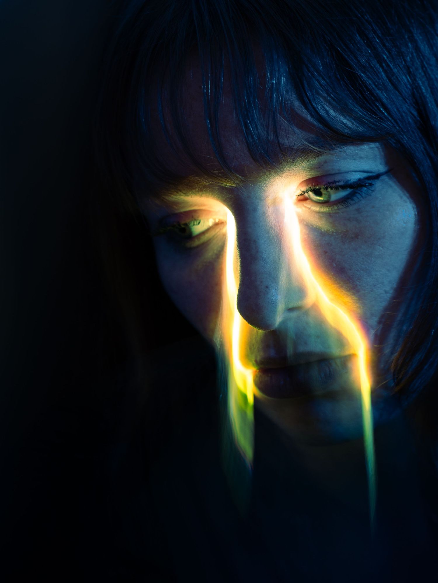 Close-up portrait of a woman with colorful light streaks across her face in a dark, moody setting.