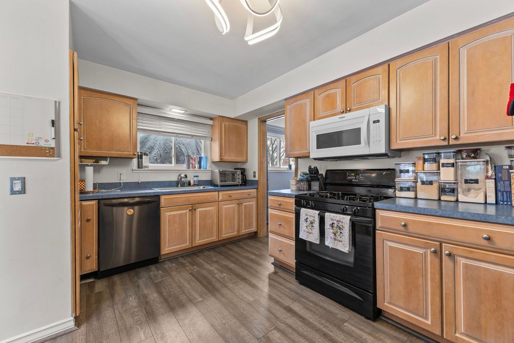 Kitchen with oak cabinets, black gas range, stainless dishwasher, white microwave, and dark countertops on hardwood floors.