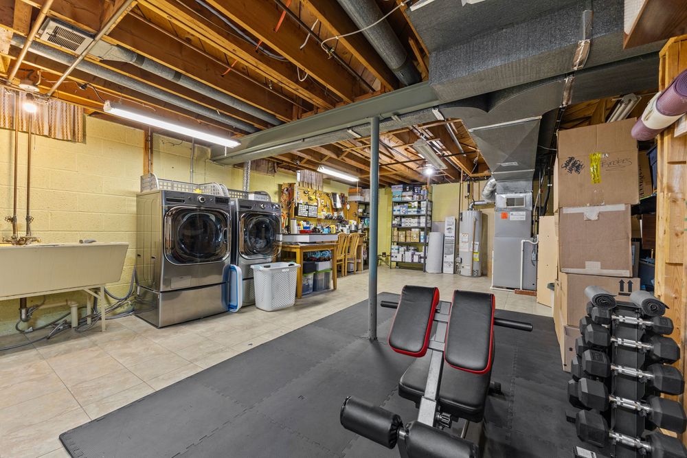 Unfinished basement with home gym equipment, washer and dryer, utility sink, and storage shelving on tile flooring.