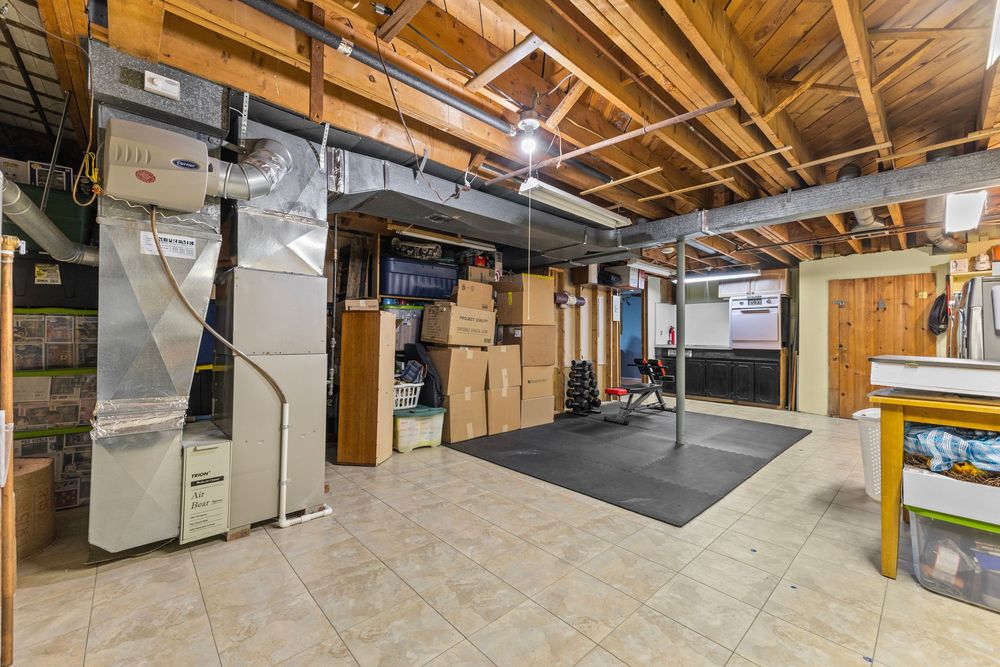 Unfinished basement with tile flooring, exposed wooden ceiling, HVAC unit, storage boxes, and a home gym workout area.