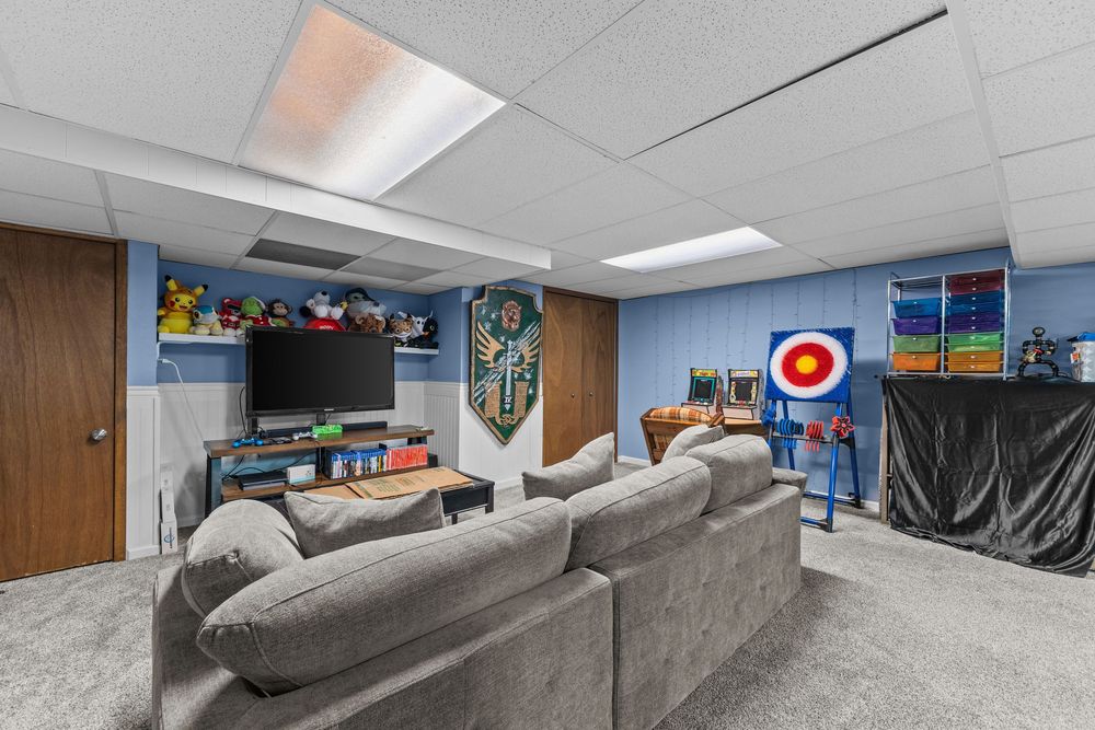 Finished basement rec room with gray sectional sofa, wall-mounted TV, blue accent walls, drop ceiling, and carpet flooring.