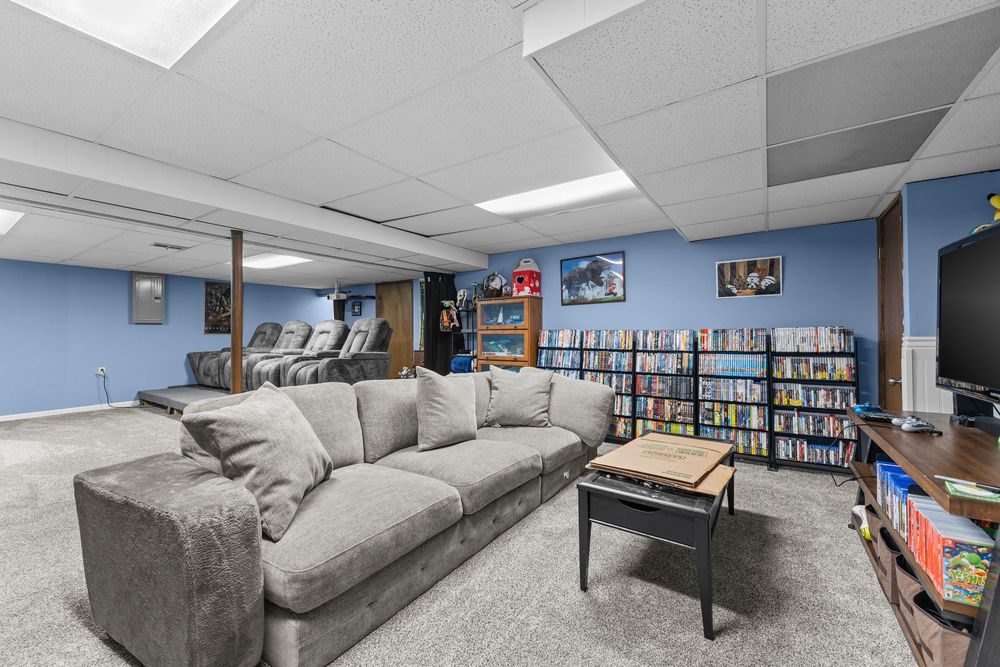Basement rec room with gray sectional sofa, blue walls, drop ceiling, carpet flooring, and large bookshelf filled with media.