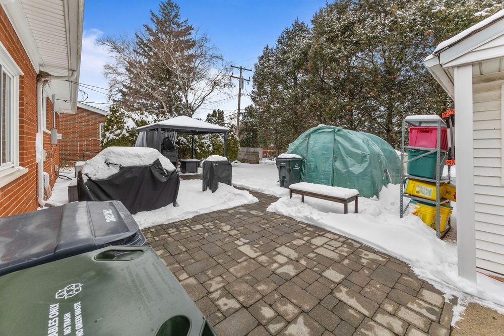 Snow-covered backyard patio with brick pavers, covered BBQ grill, outdoor furniture, and a green gazebo in winter.