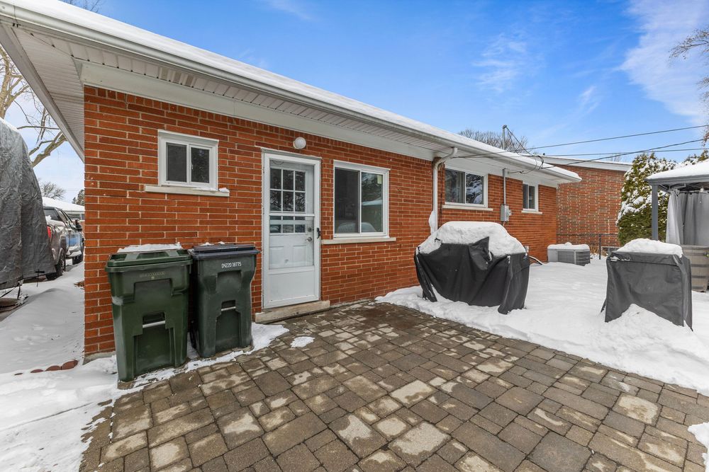 Brick bungalow exterior in winter with snow-covered patio, green garbage bins, and covered BBQ on interlocking stone driveway.