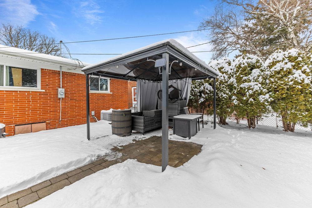 Snow-covered backyard patio with a metal gazebo structure and covered hot tub beside a brick home in winter.