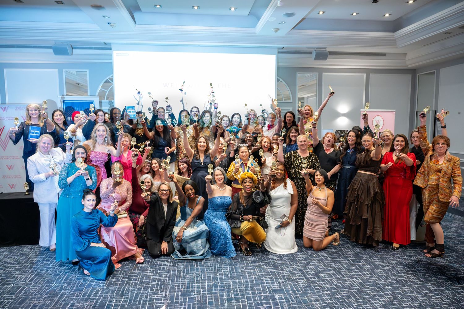 Large group of elegantly dressed women posing together at an awards ceremony in a hotel ballroom.
