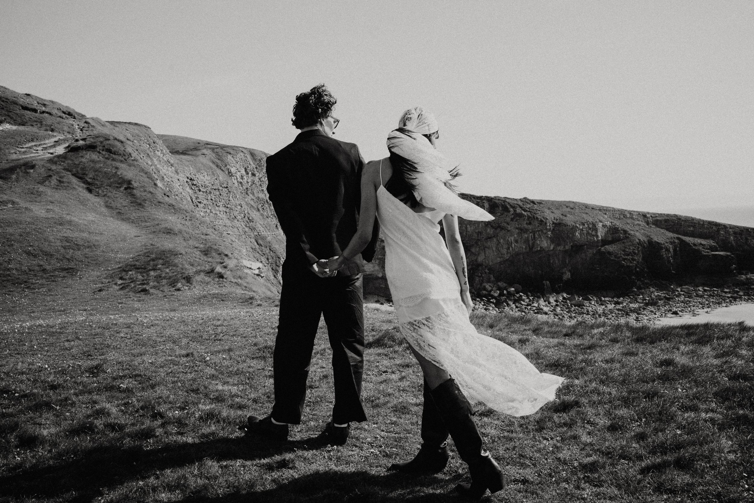 Elegant couple in wedding attire posing by white columns with ocean view, bride in lace dress shielding eyes from sun.