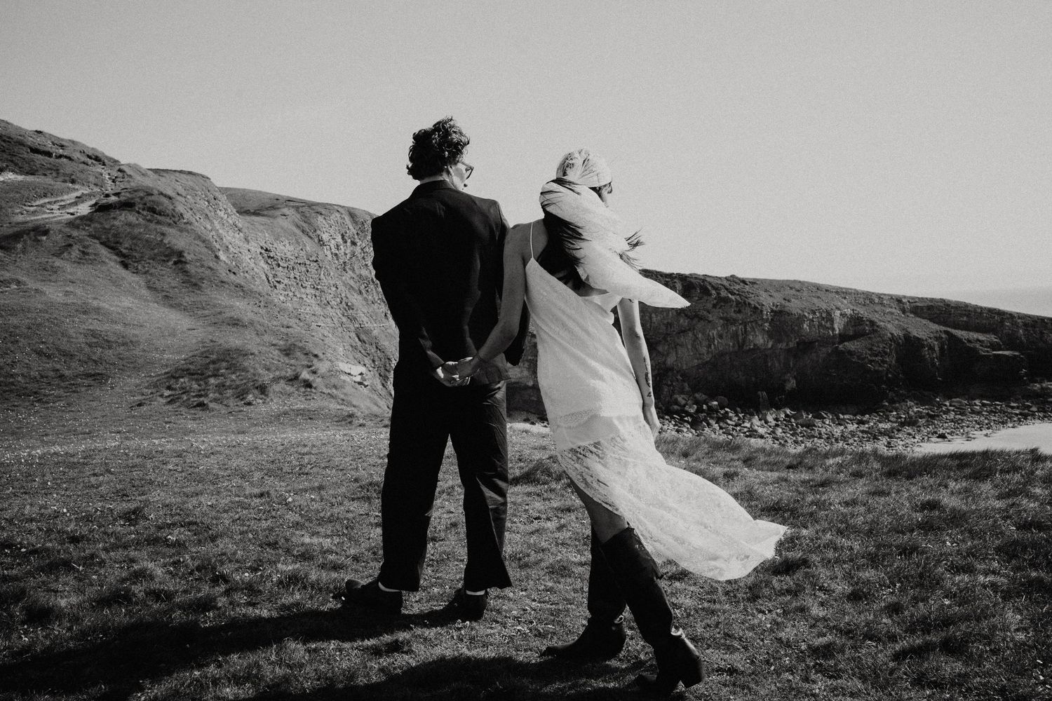 Elegant couple in wedding attire posing by white columns with ocean view, bride in lace dress shielding eyes from sun.