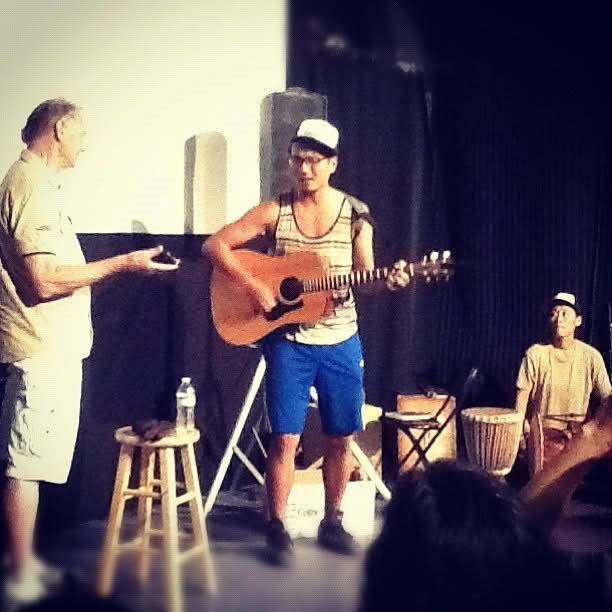 Young man playing acoustic guitar on stage with instructor and drummer performing live music.