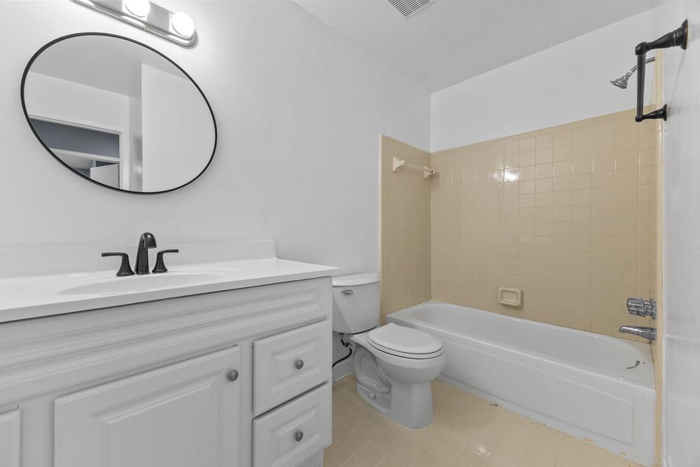 Full bathroom with white vanity, round mirror, toilet, and tiled bathtub surround.