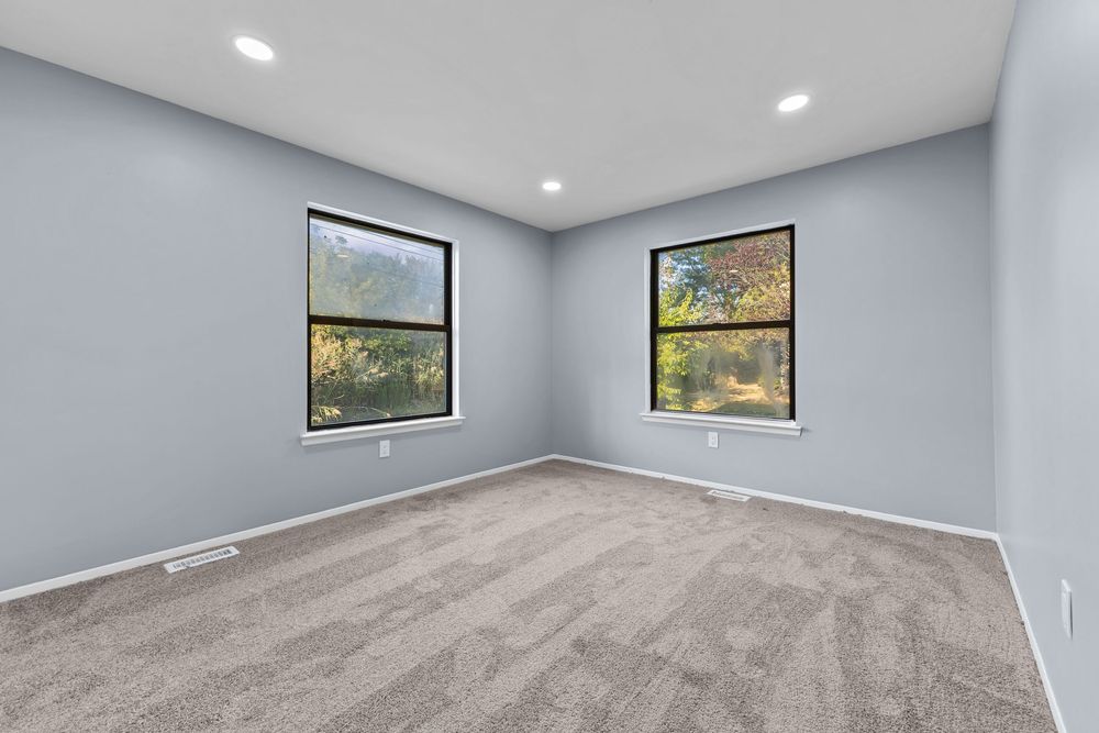 Empty bedroom with gray carpet, two windows with black frames, recessed lighting, and light blue-gray walls.