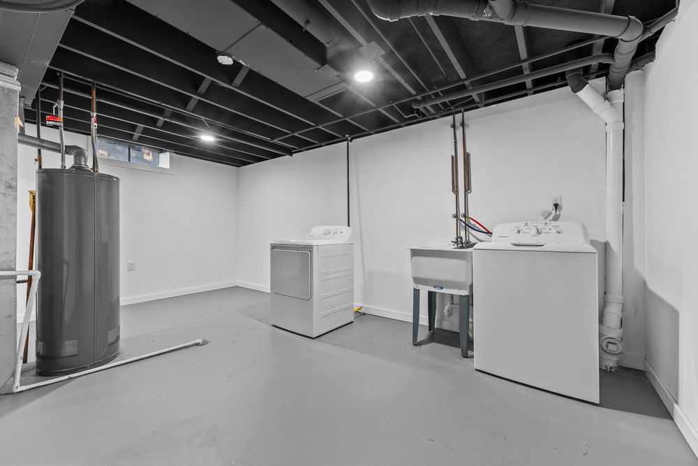 Basement laundry area with washer, dryer, utility sink, water heater, and exposed black ceiling joists.