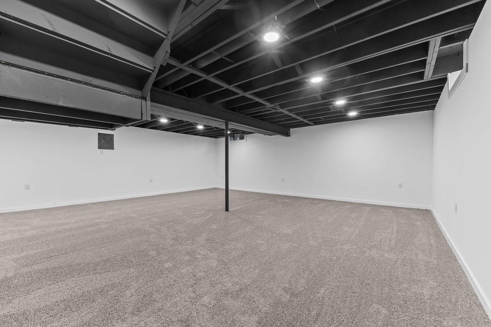 Large open basement space with gray carpet, exposed black painted ceiling joists, and recessed lighting.