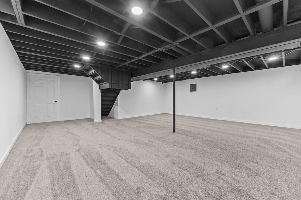 Spacious unfinished basement with gray carpet, exposed black ceiling joists, support pole, and staircase.